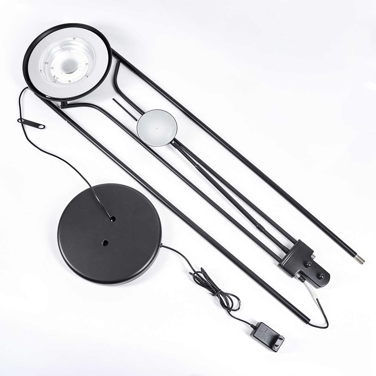 Eine zerlegte schwarze Stehlampe mit rundem Sockel, Leuchtenkopf und Netzteil, auf weißem Untergrund platziert.