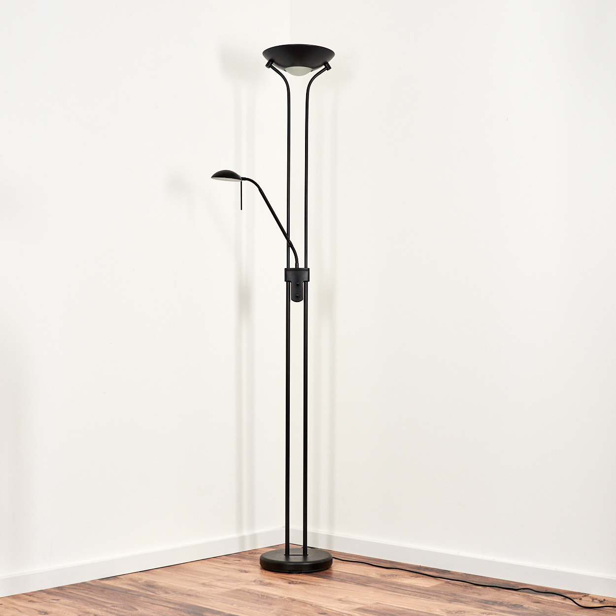 Eine schwarze Stehlampe steht in einer Ecke. Die Lampe hat einen kreisförmigen Fuß, einen hohen vertikalen Schaft und zwei Leuchten.