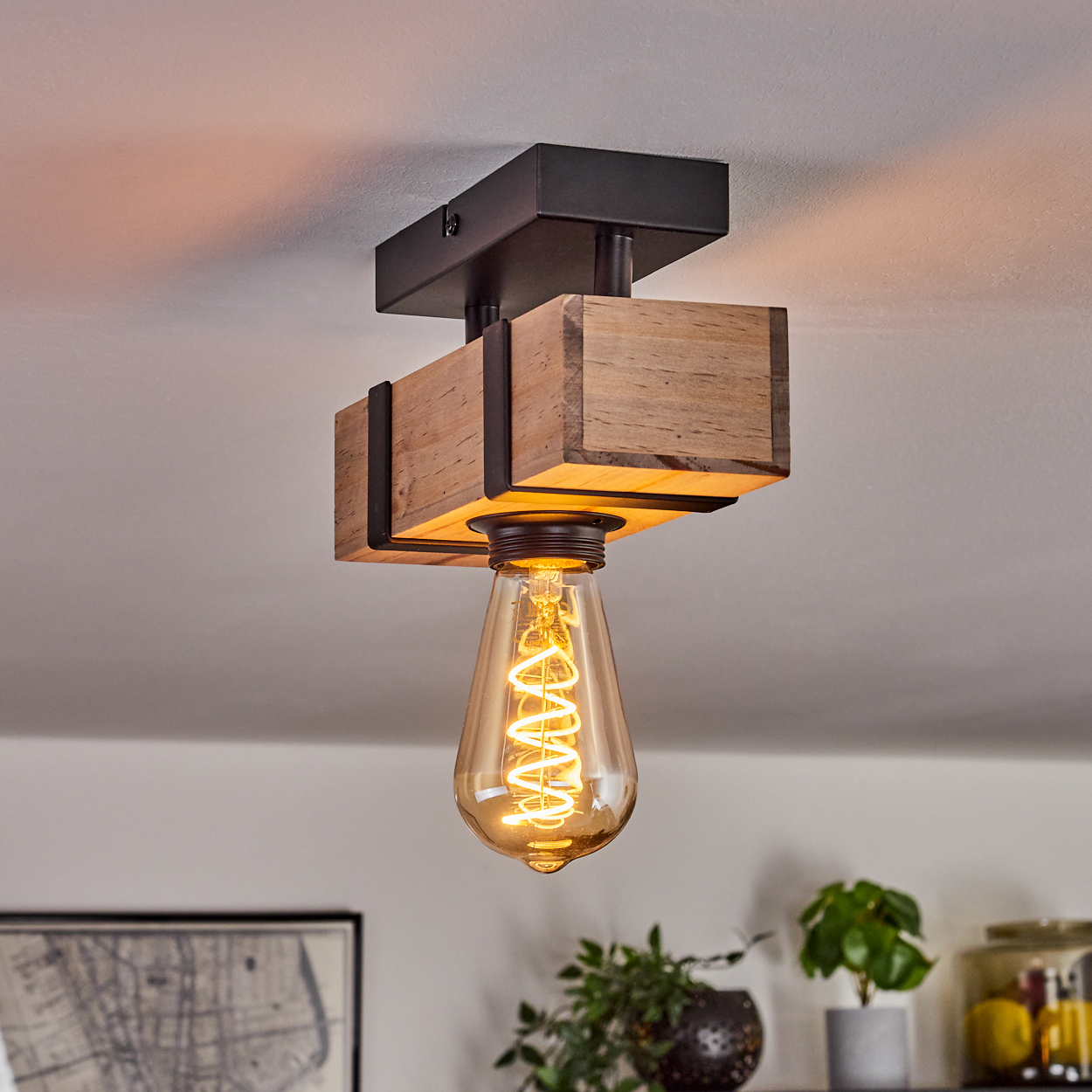 Deckenlampe mit Holzbalken und großer Glühbirne mit Wendelfaden. Schwarze Metallakzente, weiße Decke, verschwommener Hintergrund mit Pflanzen.