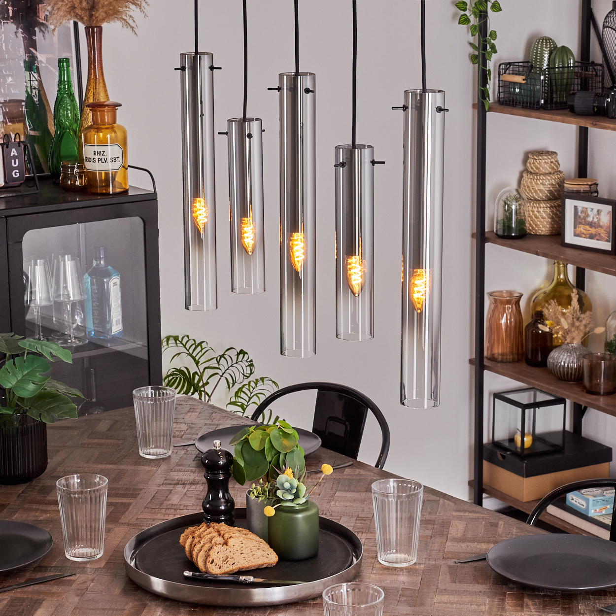 Ein Esstisch ist mit Tellern, Gläsern und einem Mittelstück gedeckt. Darüber hängen zylindrische Lichter. Ein Schrank und Regale sind im Hintergrund.
