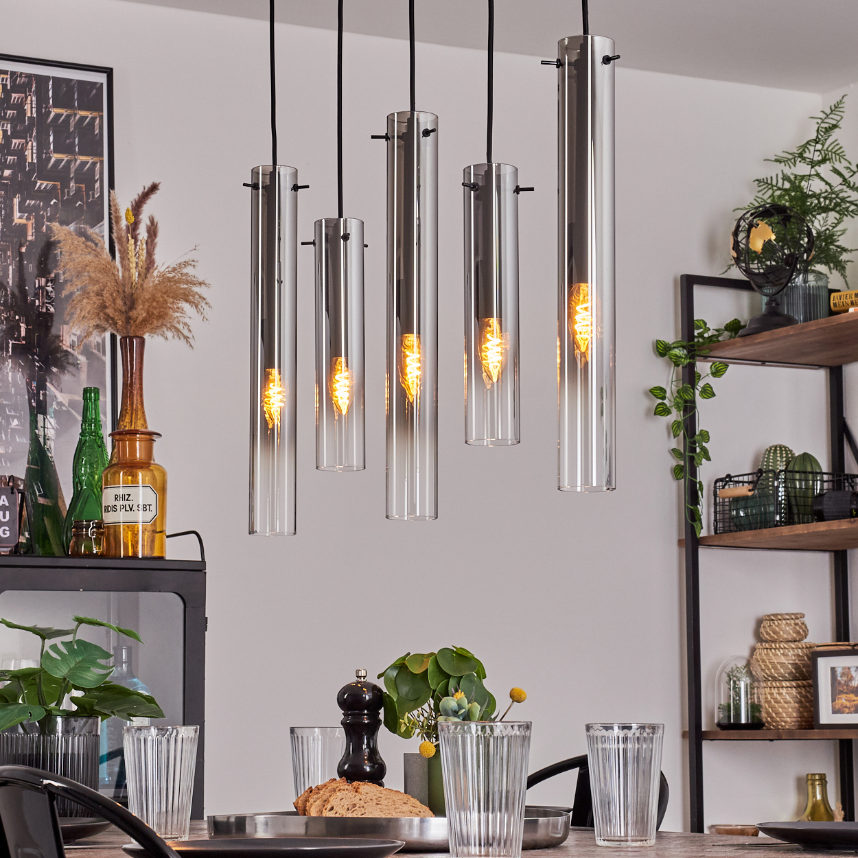 Esszimmer mit Hängelampen. Tisch mit Gläsern, Brot und Pfeffermühle gedeckt. Regale mit Pflanzen und Dekoration.