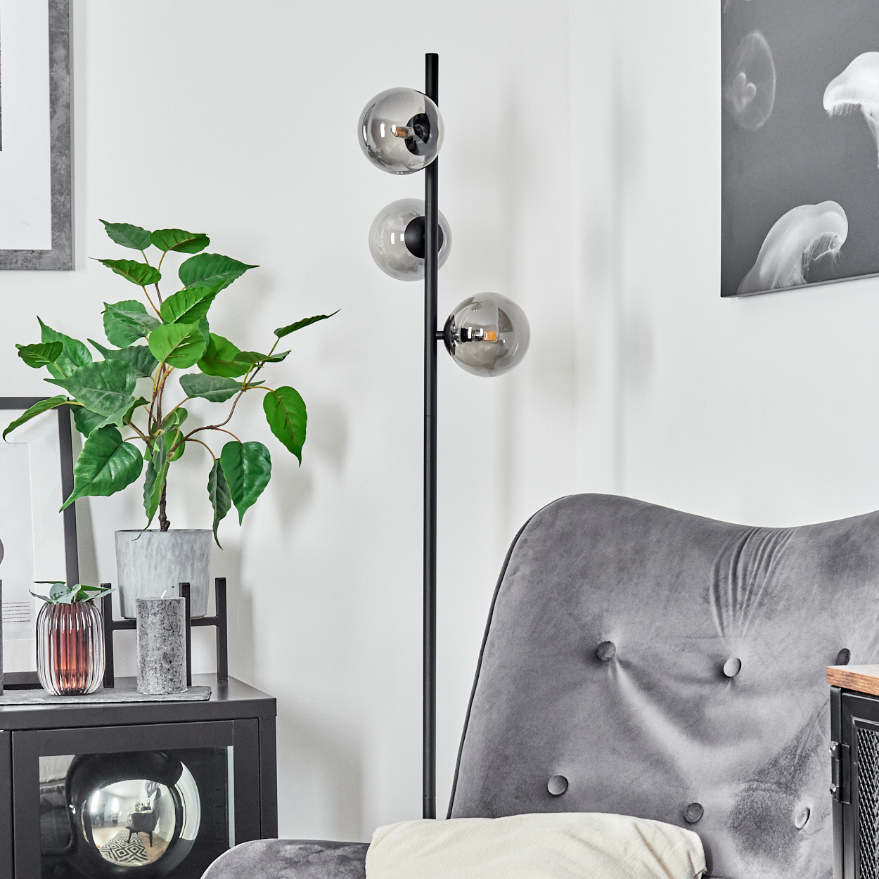 Ein modernes Wohnzimmer mit grauem Sessel, Stehlampe mit drei Glaskugeln und Topfpflanze. Kunstwerke schmücken die Wände.