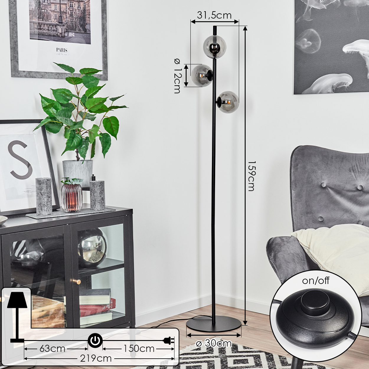 Eine schwarze Stehlampe mit drei Glaskugeln. Sie steht neben einem Schrank, einem grauen Sessel und einem Teppich. Die Lampe ist eingeschaltet.