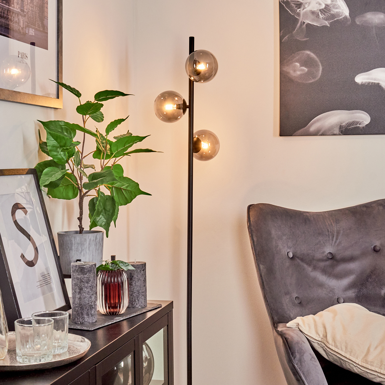 Ein Raum mit einer Stehlampe, einer Pflanze und einem gerahmten Bild. Im Hintergrund steht ein grauer Sessel.