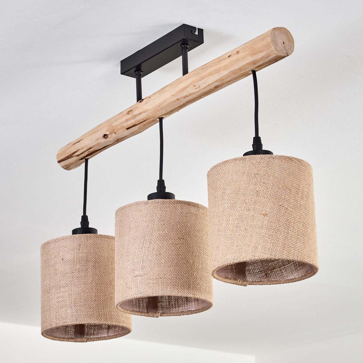 Ein Holzbalken mit drei Pendelleuchten mit Juteschirmen hängt von einer weißen Decke. Die Leuchte hat schwarze Kabel und Halterungen.