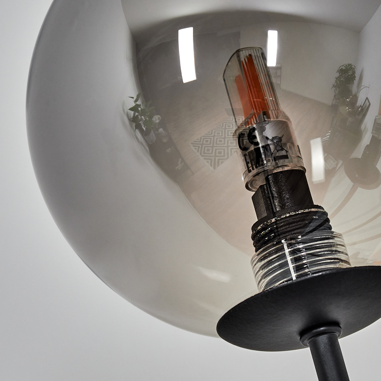 Nahaufnahme einer modernen Lampe mit Glaskolben und schwarzem Sockel. Die Glühbirne spiegelt einen Raum mit Pflanzen und geometrischen Mustern wider.