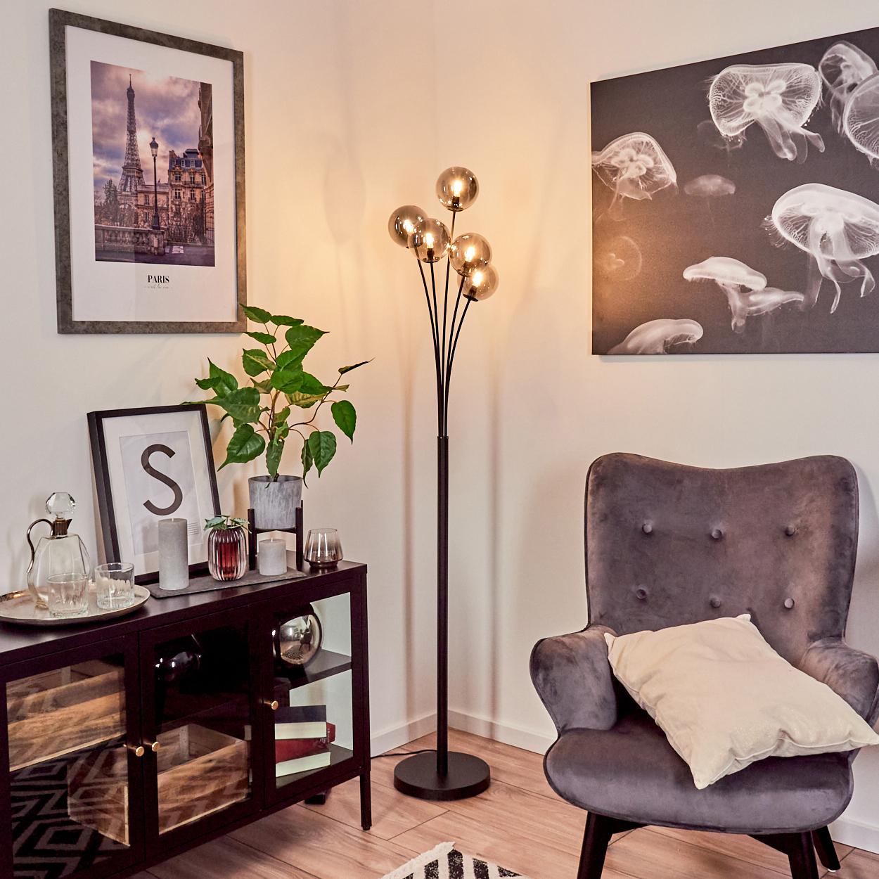 Ein Wohnzimmer mit Stehlampe, Kunst und grauem Sessel. Ein Schrank beherbergt Deko und eine Pflanze. Die Szene ist hell und gemütlich.
