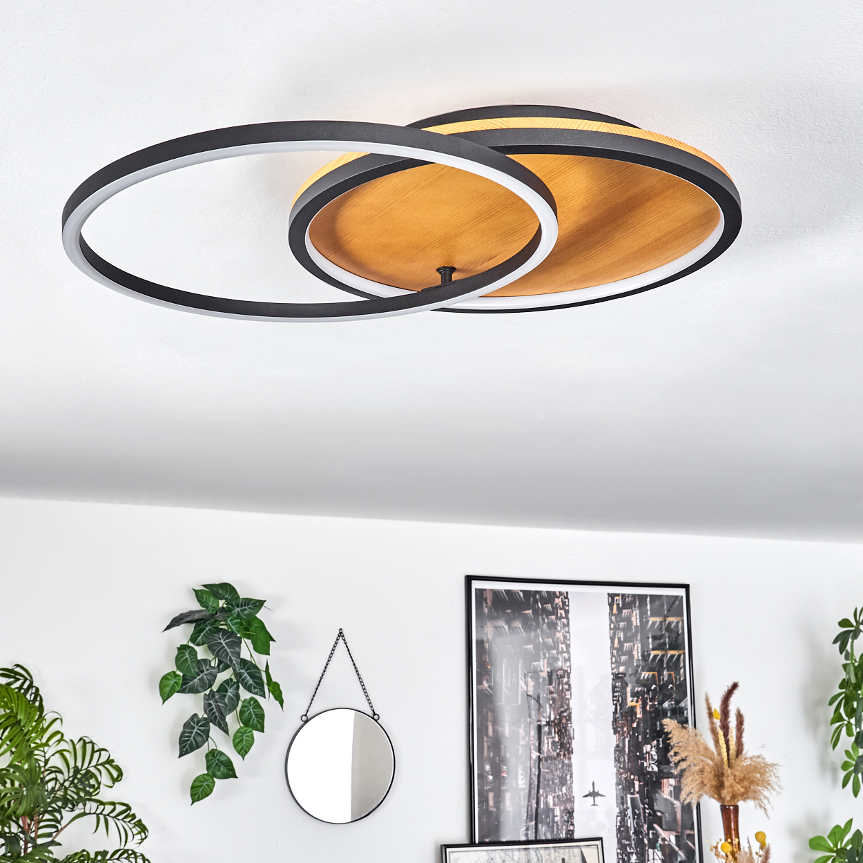 Eine Deckenleuchte mit schwarzen Ringen und Holzsockel. An der weißen Wand befinden sich ein Spiegel und ein Stadtbildkunstwerk. Pflanzen sind an den Seiten.