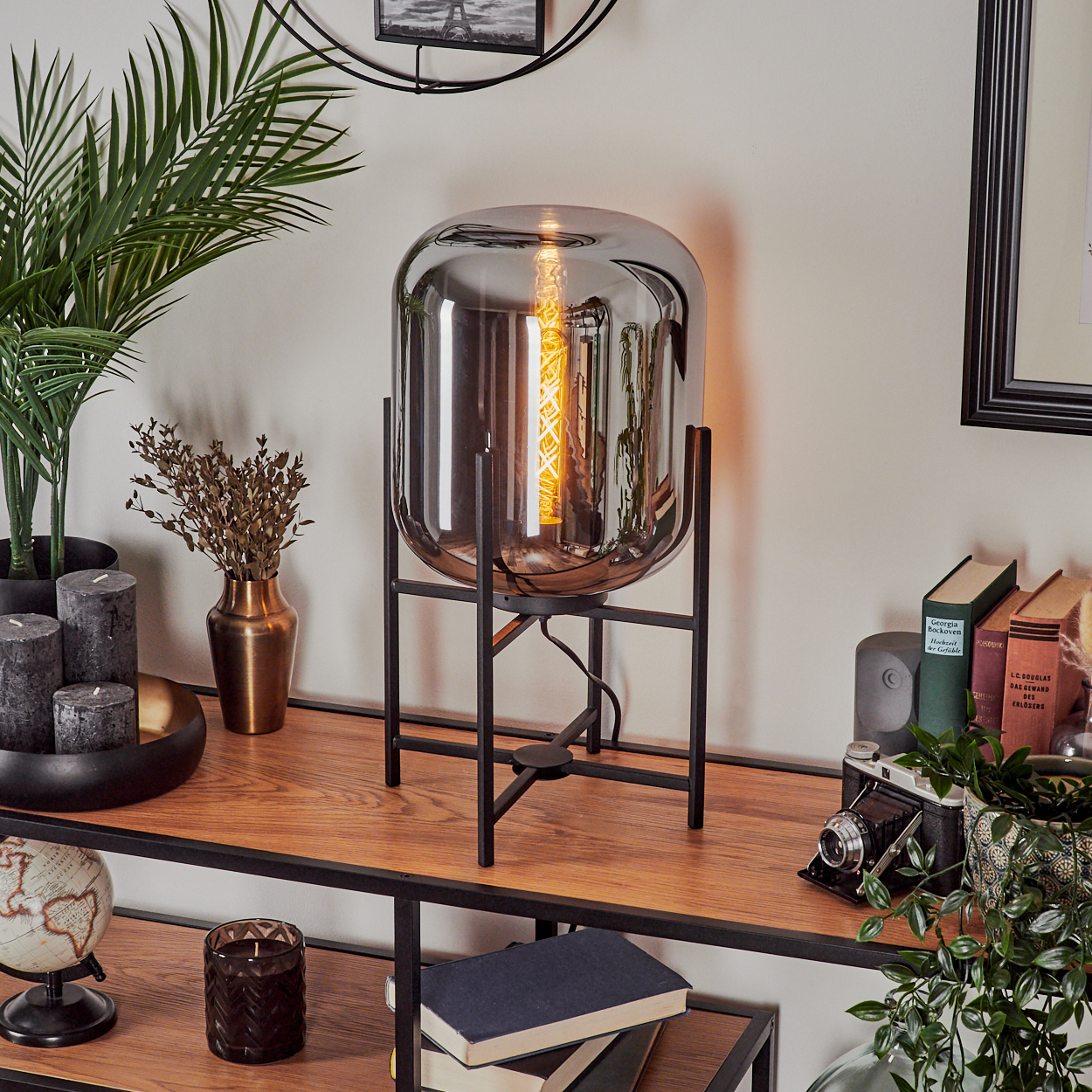 Eine moderne Lampe mit Glasschirm auf einem Holzregal. Es gibt Bücher, Pflanzen, Kerzen und einen Globus, was eine gemütliche Atmosphäre schafft.