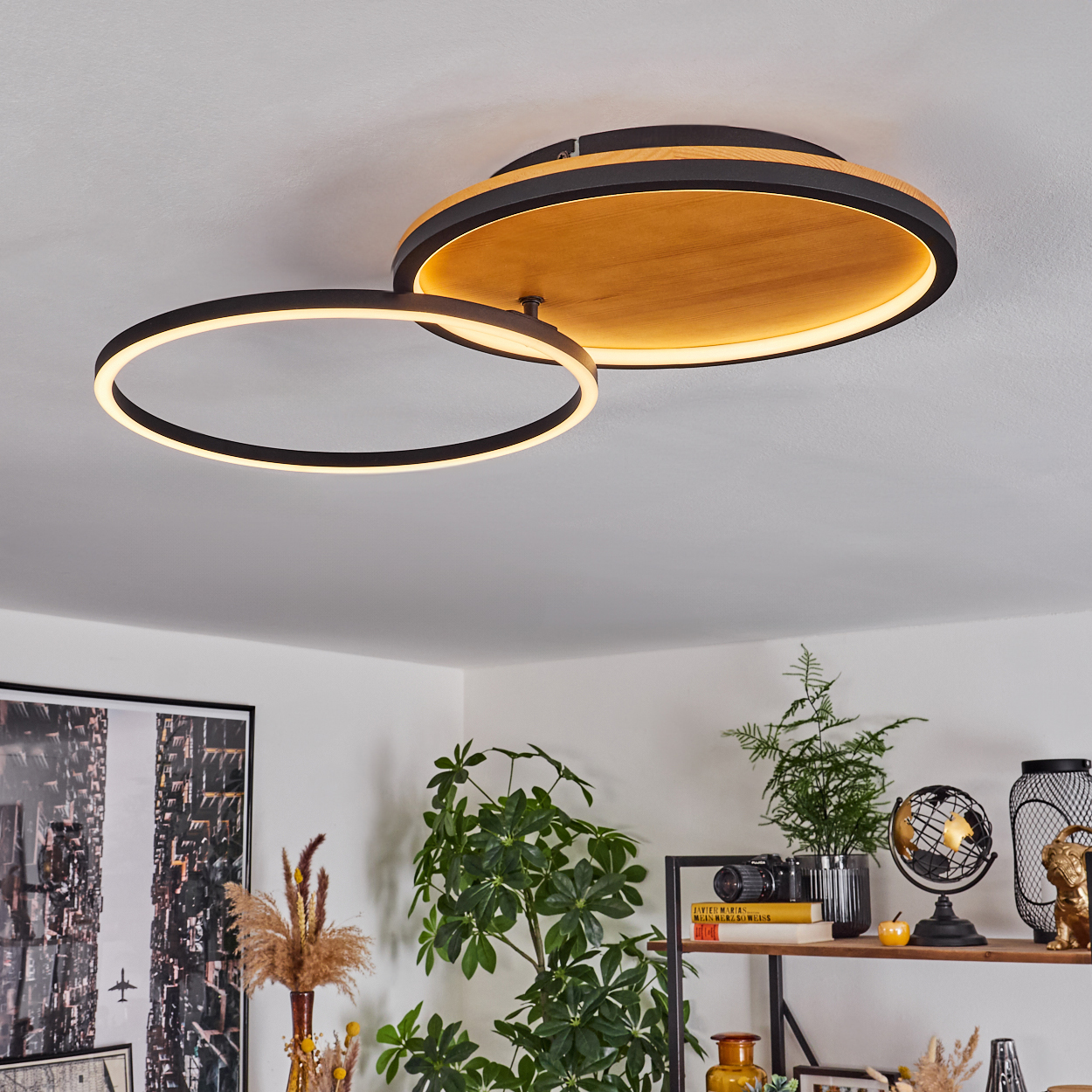 Moderne Deckenleuchte mit zwei miteinander verbundenen Leuchtringen. Holz- und schwarzes Design. Raum mit Pflanzen und Dekoration im Hintergrund.