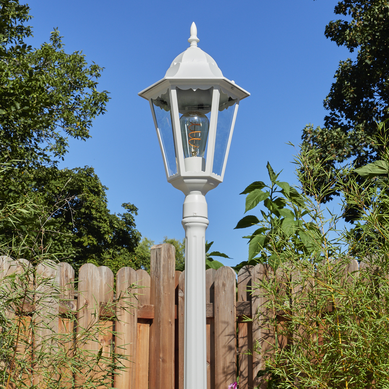 Weiße Gartenlampe mit leuchtender Glühbirne, Holzzaun und grüne Pflanzen vor blauem Himmel.