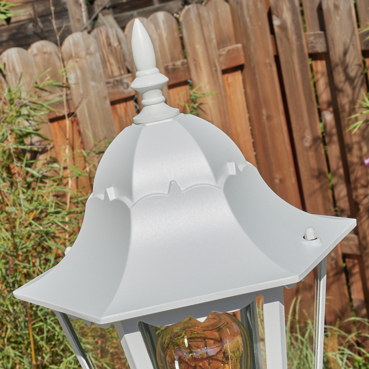 Eine weiße Außenlampe mit dekorativer Spitze. Im Hintergrund ein Holzzaun und grüne Pflanzen.
