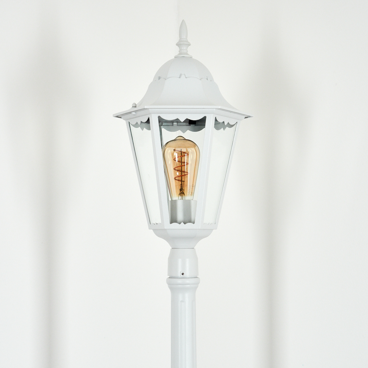 Eine weiße Außenlampe mit einer Vintage-Glühbirne. Sie steht vor einer weißen Wand. Die Lampe hat eine dekorative Spitze und klares Glas.