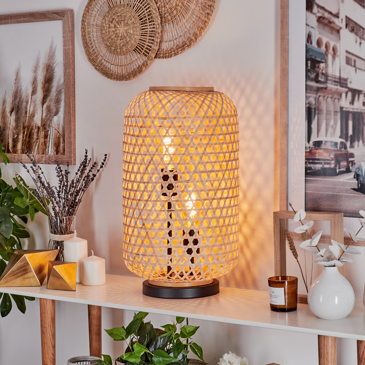 Eine Bambuslampe leuchtet auf einem weißen Tisch. Dahinter befinden sich gerahmte Kunstwerke und Dekorationen. Pflanzen und Kerzen sind ebenfalls vorhanden.
