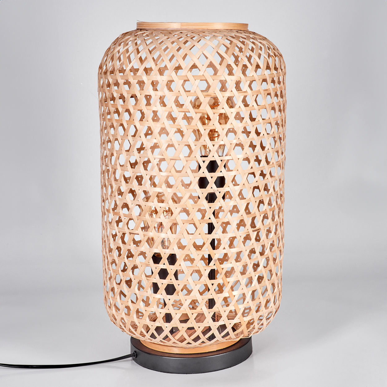 Eine gewebte Bambuslampe steht auf einem dunklen Sockel mit einem Lichtkabel. Sie hat eine zylindrische Form und eine Glühbirne im Inneren.