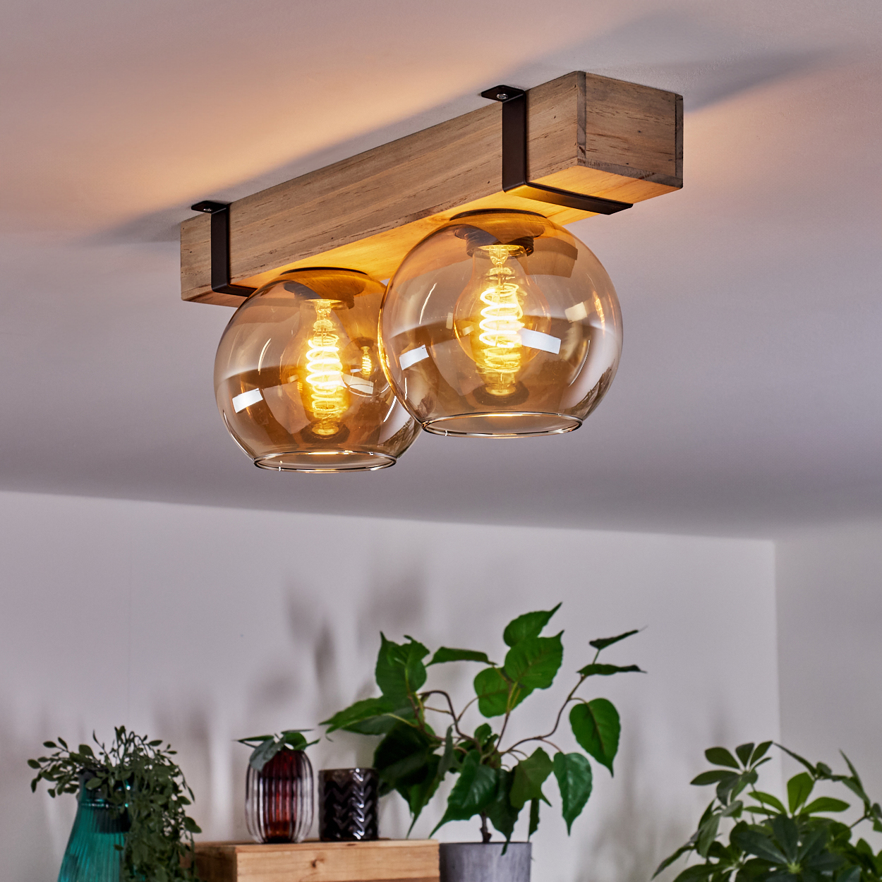Deckenleuchte mit zwei Glaskugeln und freiliegenden Glühbirnen. Holzbalken und dekorative Pflanzen im Vordergrund. Warme Beleuchtung.