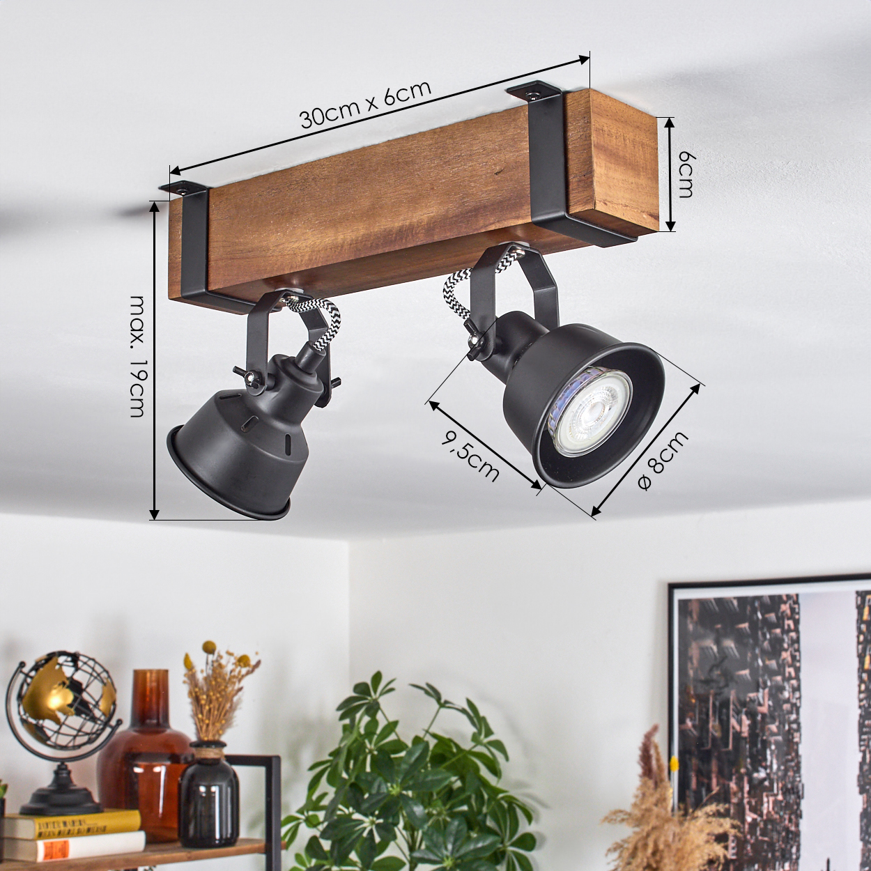 Ein Holzbalken mit zwei schwarzen Strahlern ist an einer weißen Decke montiert. Abmessungen sind angegeben. Der Hintergrund zeigt ein Regal mit Deko.