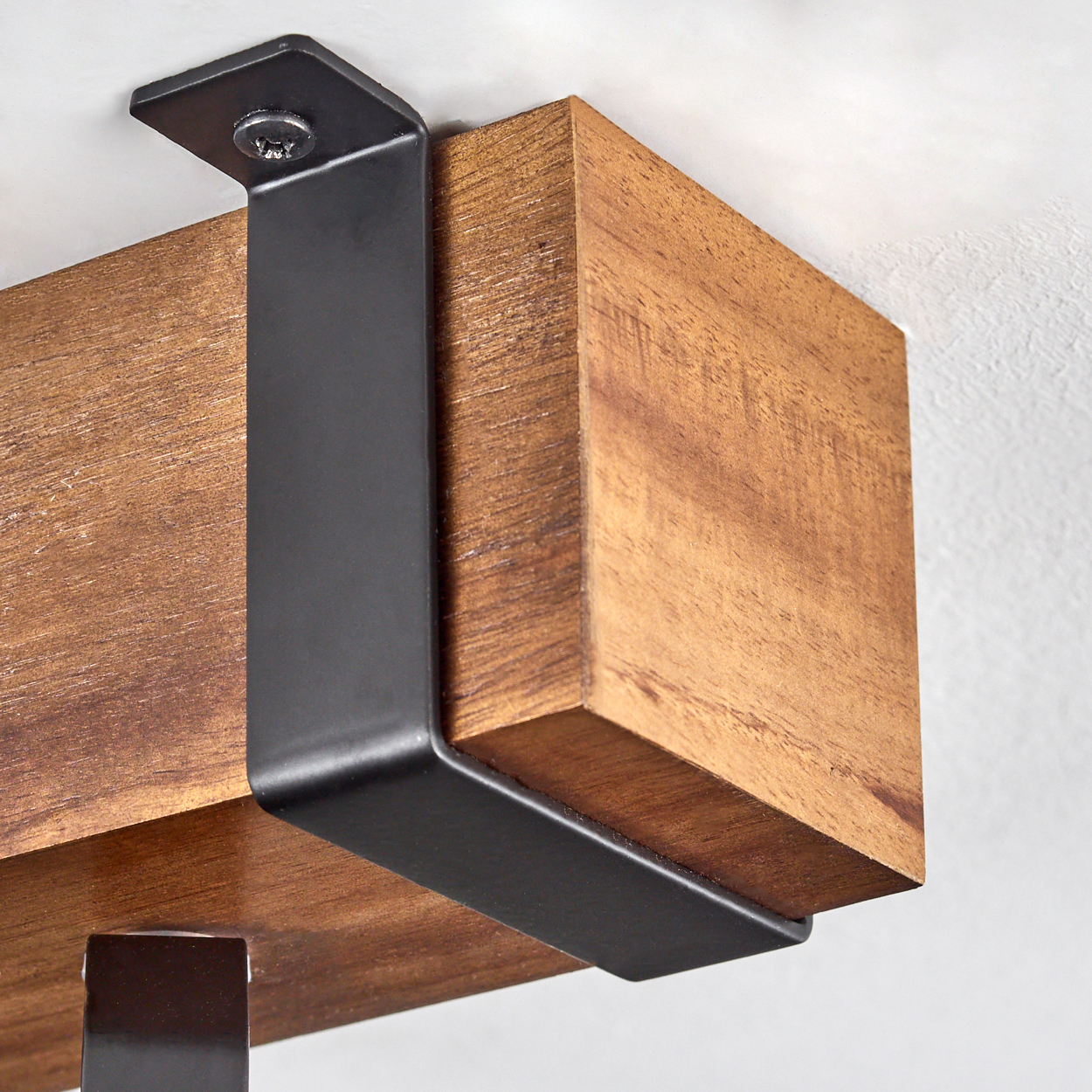 Ein Holzbalken, der von einer schwarzen Metallhalterung getragen wird und an einer weißen Wand befestigt ist. Das Holz ist braun und die Halterung dunkel.