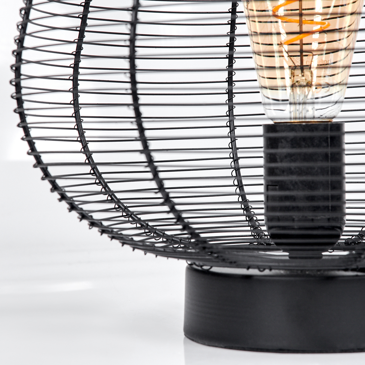 Eine schwarze Drahtlampe mit einer Glühbirne. Die Glühbirne hat einen spiralförmigen Glühfaden. Die Lampe steht auf einem runden schwarzen Sockel. Weißer Hintergrund.