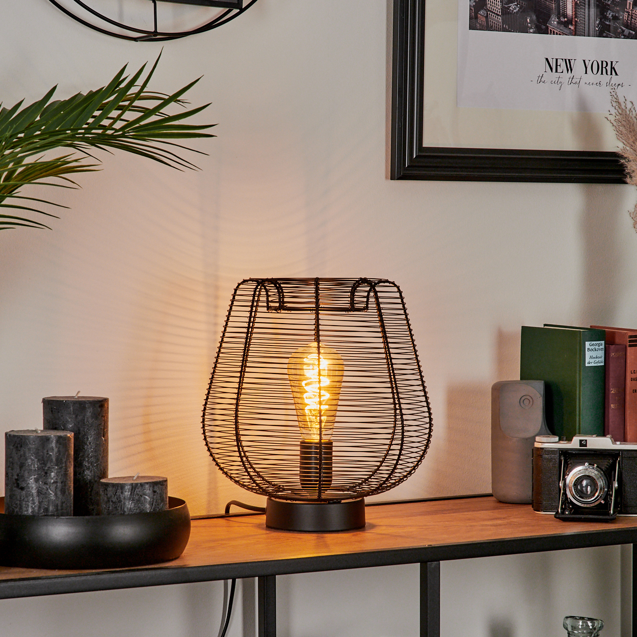 Eine schwarze Drahtgitterlampe mit leuchtender Glühbirne steht auf einem Holzregal. Kerzen, Bücher und eine Kamera befinden sich ebenfalls auf dem Regal.