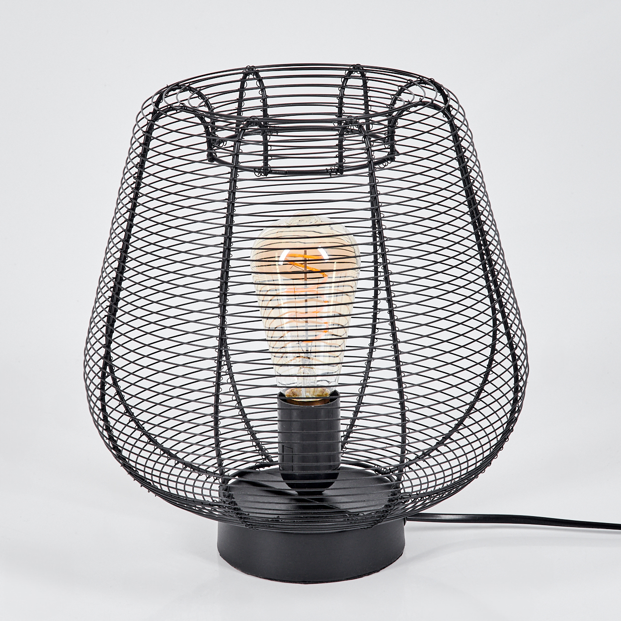 Eine schwarze Drahtlampe mit einer Glühbirne im Inneren. Die Lampe steht auf einer weißen Oberfläche, mit einem schwarzen Kabel.
