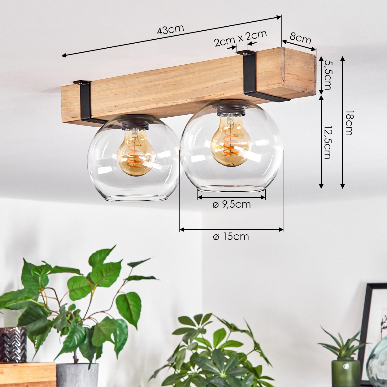 Eine Deckenlampe aus Holzbalken mit zwei Glaskugeln, die jeweils eine leuchtende Glühbirne enthalten. Pflanzen im Vordergrund.