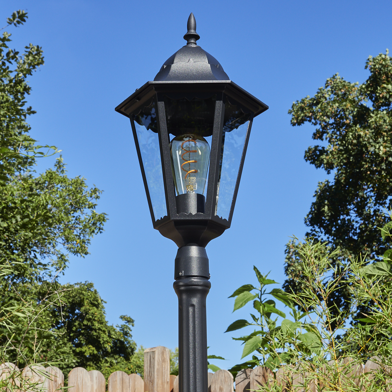Eine schwarze Außenlampe mit dekorativer Glühbirne steht vor blauem Himmel, umgeben von Bäumen und einem Holzzaun.