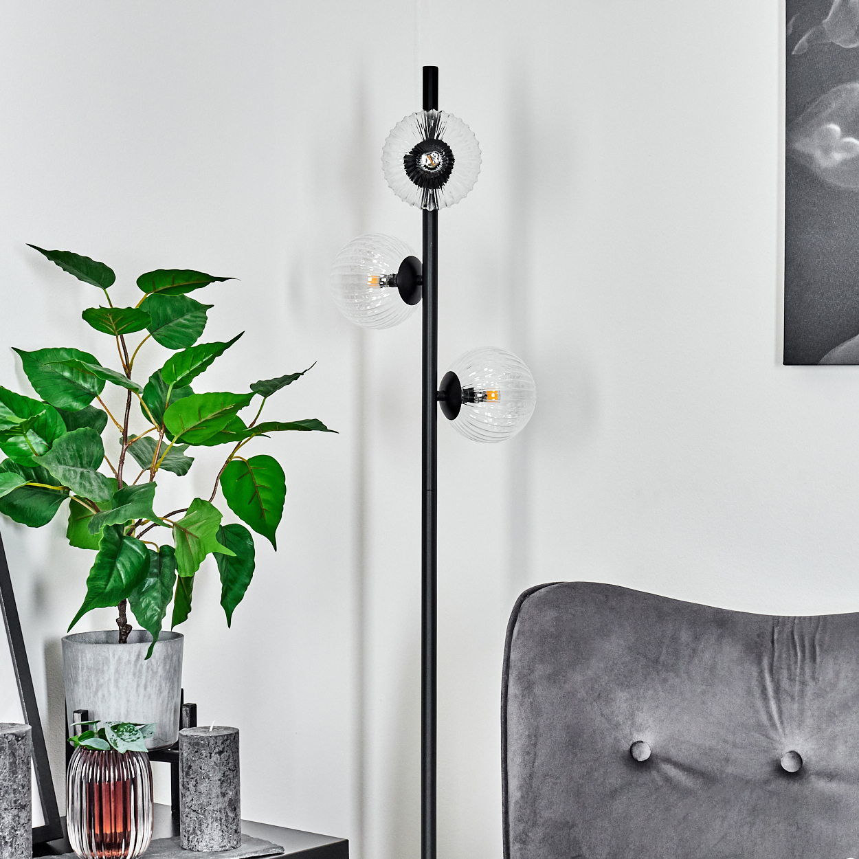 Modernes Interieur mit schwarzer Stehlampe, Topfpflanze, Kerzen und grauem Sessel. Die Lampe hat drei Glaskugelleuchten.