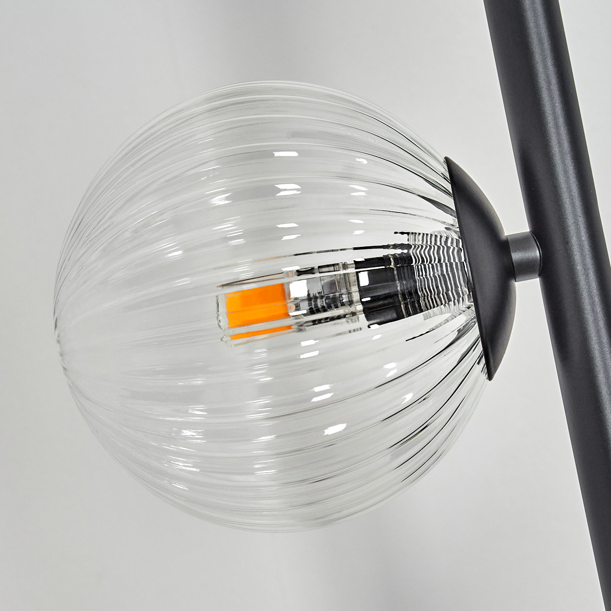Nahaufnahme einer durchsichtigen, geriffelten Glaskugel mit orangefarbenem Licht im Inneren. Von schwarzem Metallrahmen getragen, vor weißem Hintergrund.