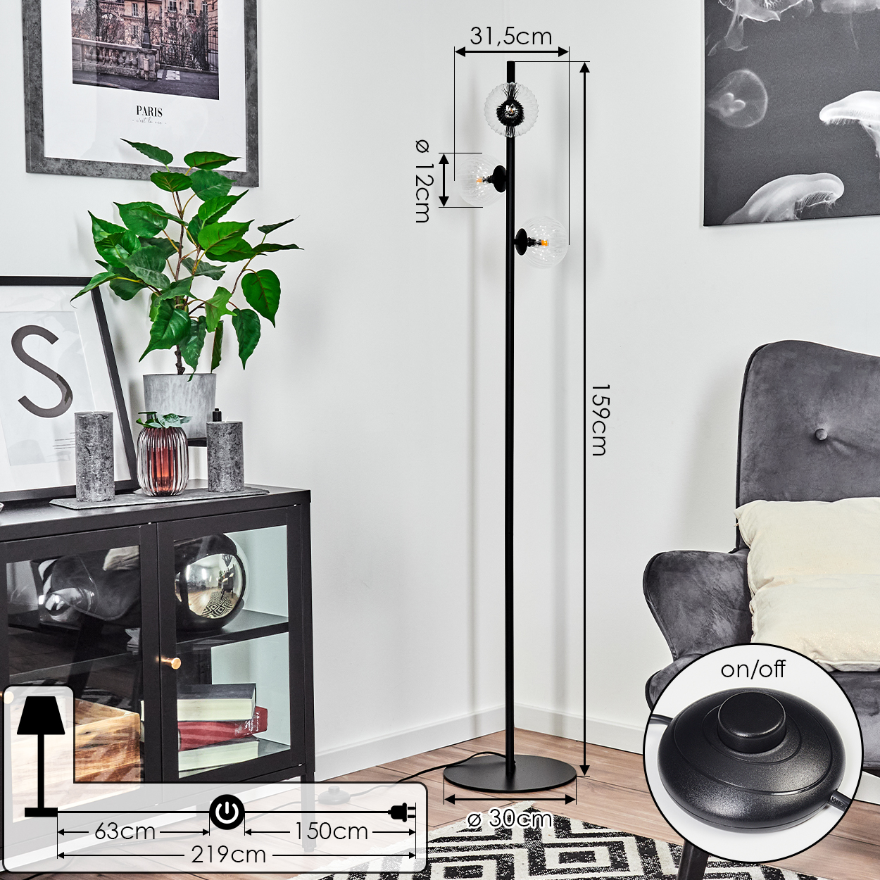 Eine schwarze Stehlampe mit drei Glaskugelleuchten. Im Hintergrund ein schwarzer Schrank mit Dekoration und ein grauer Sessel.