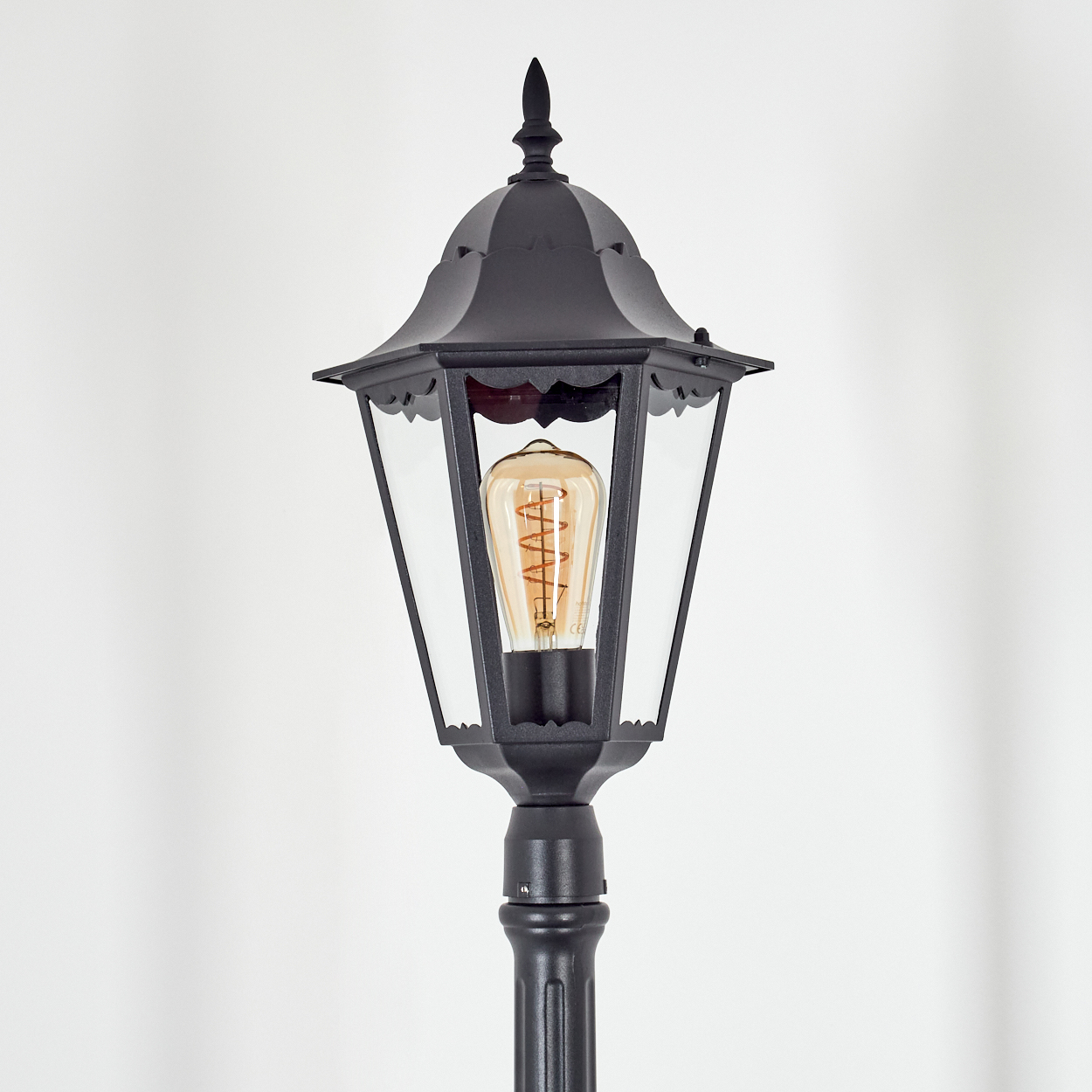 Eine schwarze Vintage-Straßenlaterne mit dekorativer Spitze, Glasscheiben und einer warm getönten Glühbirne vor weißem Hintergrund.