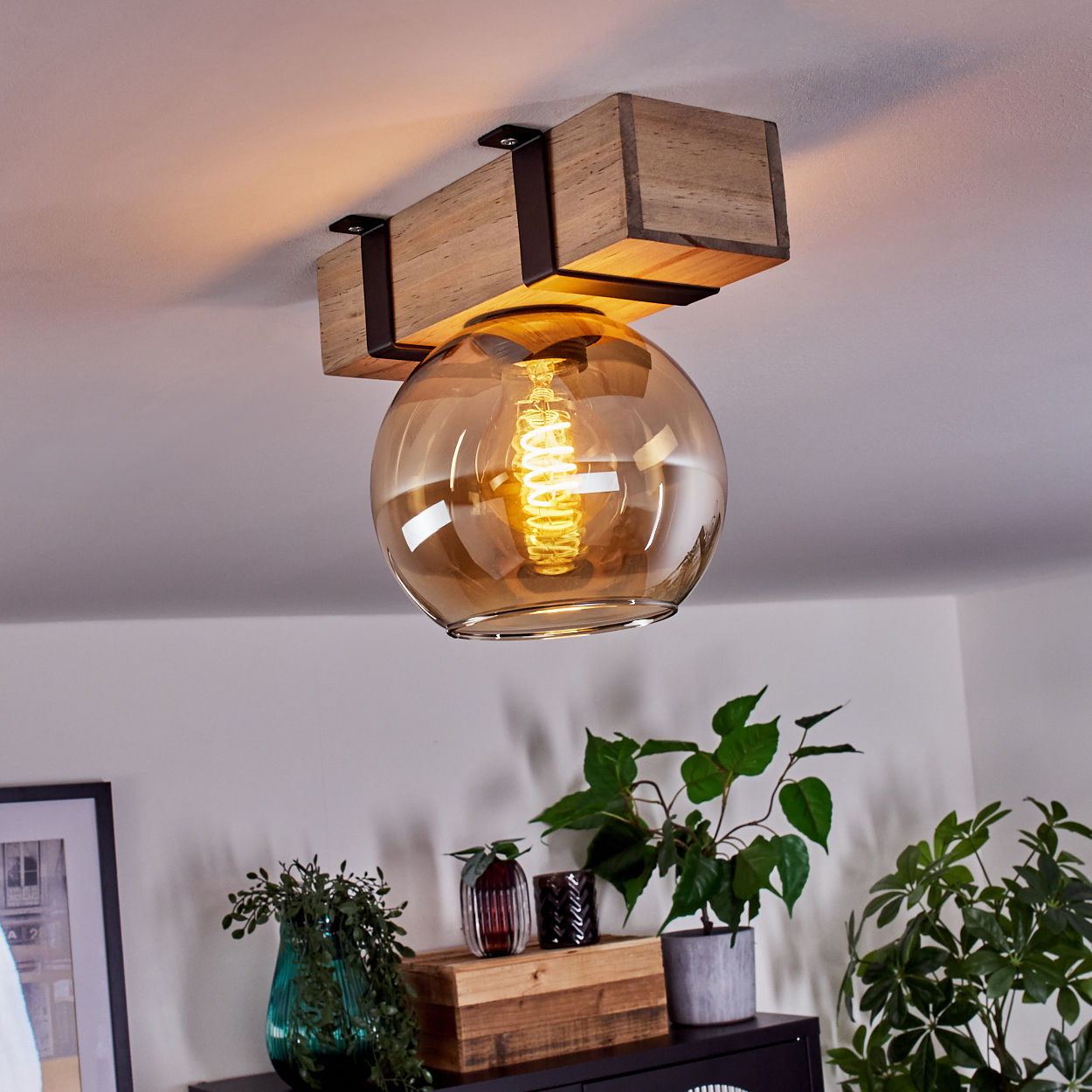 Eine Deckenlampe mit Holzbalken, Glaskugel und sichtbarer Glühbirne. Darunter Pflanzen und Dekoration auf schwarzer Oberfläche.