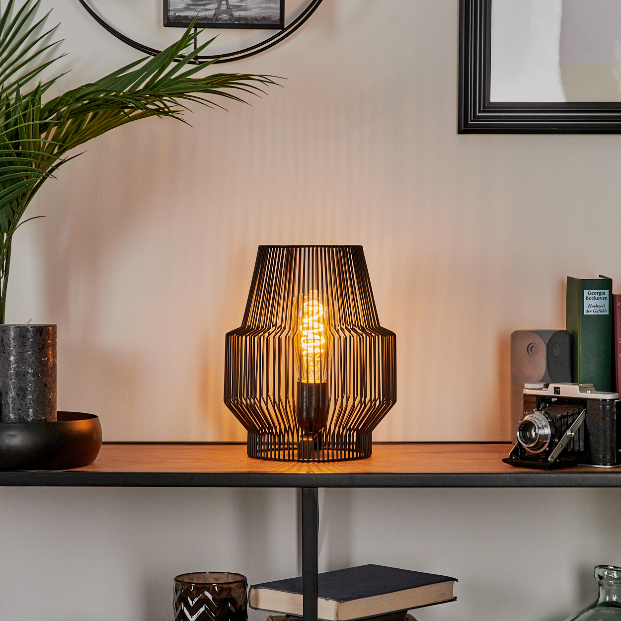 Eine schwarze Lampe mit leuchtender Glühbirne steht auf einem Regal mit Büchern und einer Kamera. Links sind eine Pflanze und eine Kerze.