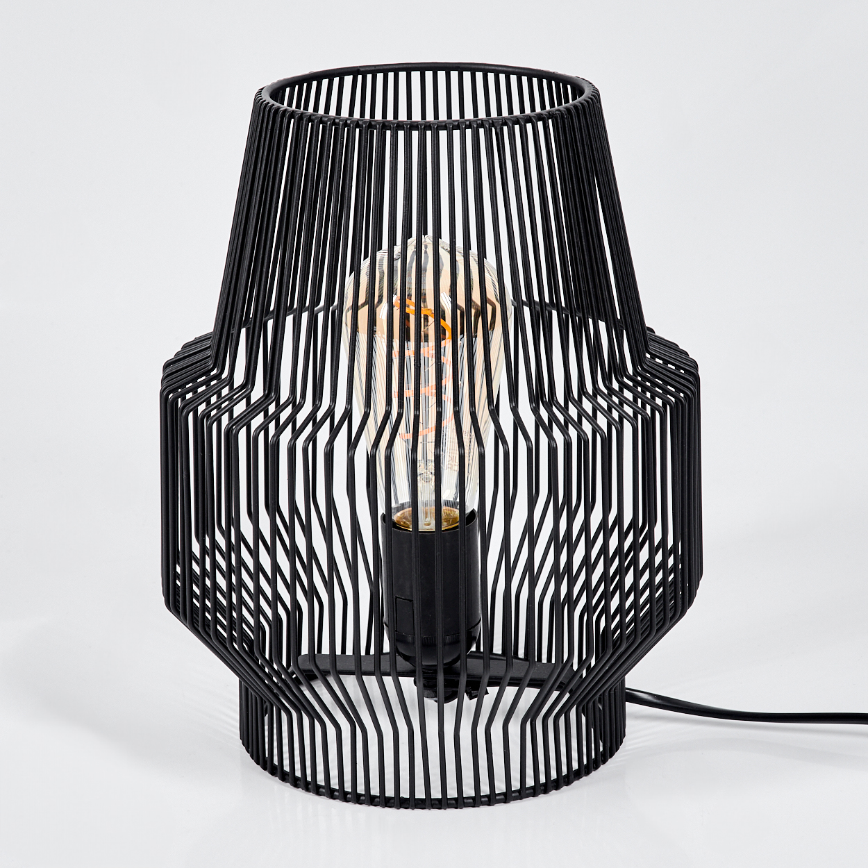 Schwarze Metallkäfiglampe mit Glühbirne. Die Lampe steht auf einer weißen Oberfläche mit schwarzem Kabel.