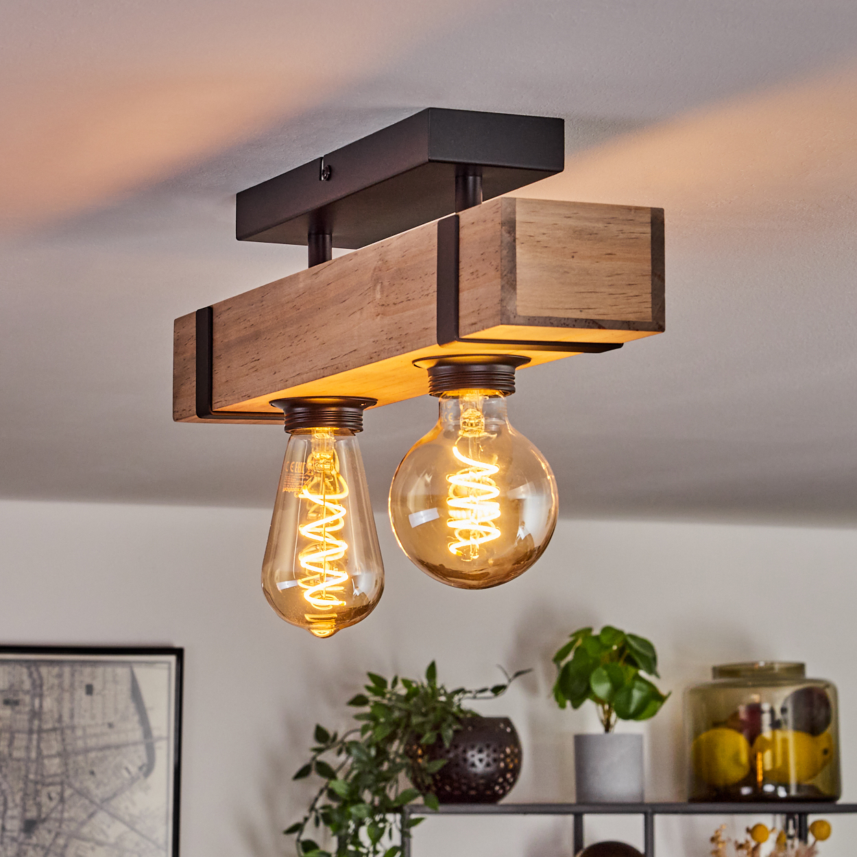 Eine Deckenlampe aus Holzbalken mit zwei leuchtenden Glühbirnen. Dekorative Pflanzen und ein Glas mit Zitronen stehen auf einem Regal darunter.