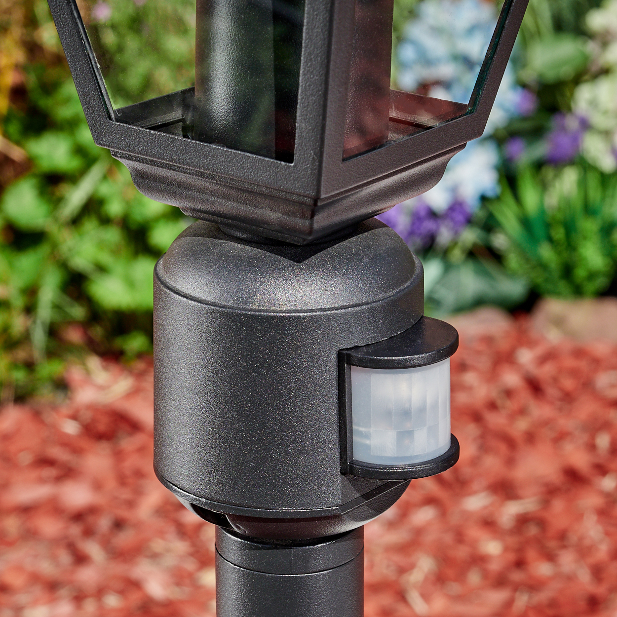 Eine schwarze Außenlampe mit Bewegungssensor. Sie steht auf einem Pfahl mit roter Mulch im Hintergrund und Grün.