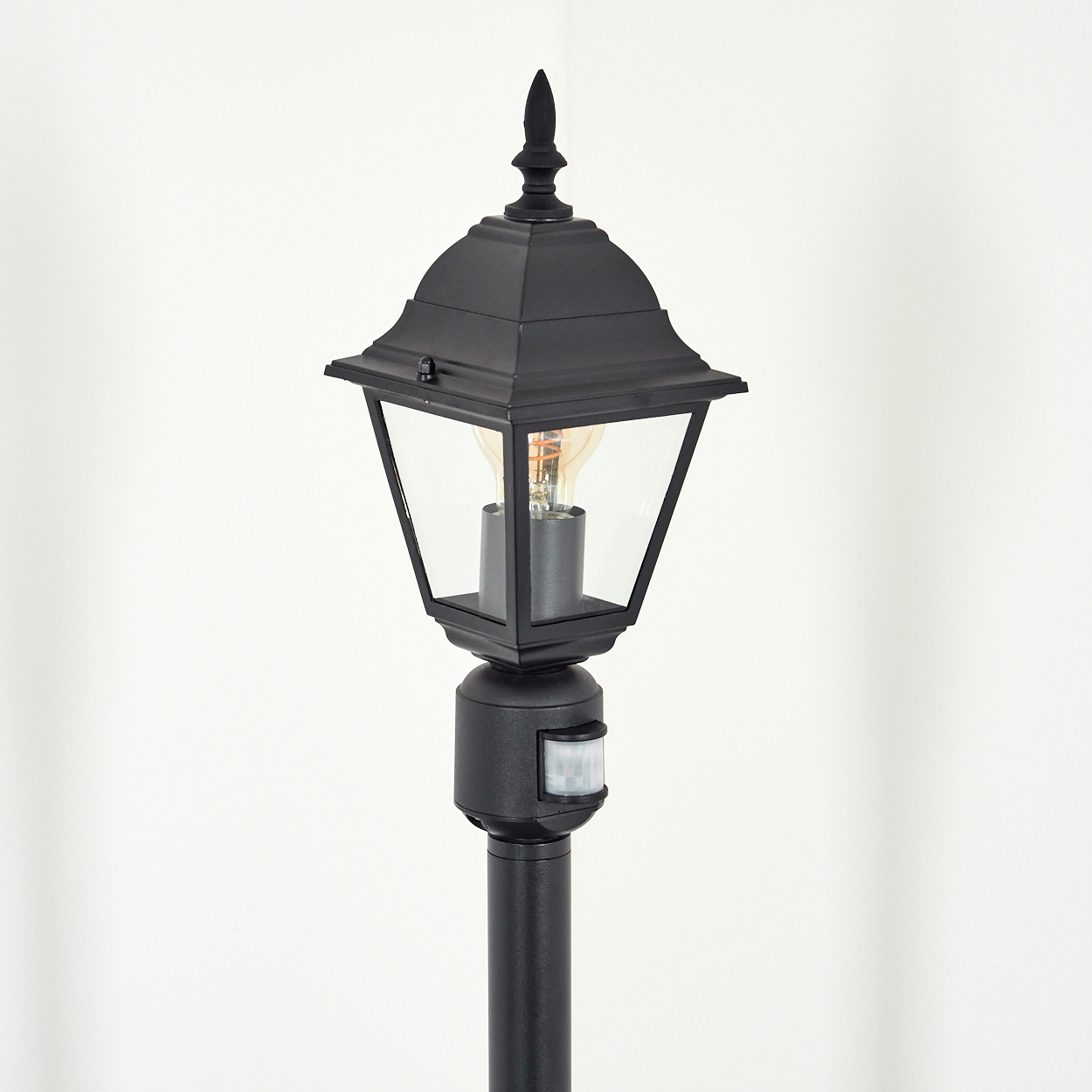 Schwarze Außenlampe mit Glaseinfassung, Glühbirne und Bewegungssensor. Der Hintergrund ist weiß.