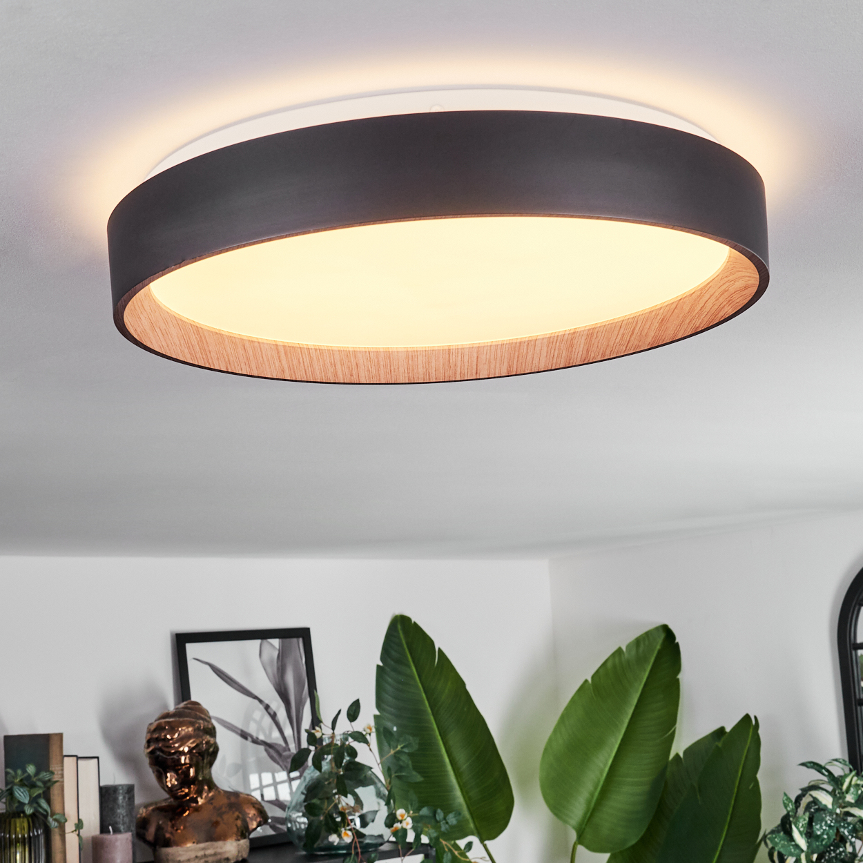 Eine moderne Deckenlampe mit einem schwarzen Außenring und einem inneren Ring aus Holz. Das Zimmer hat Pflanzen und Kunst.