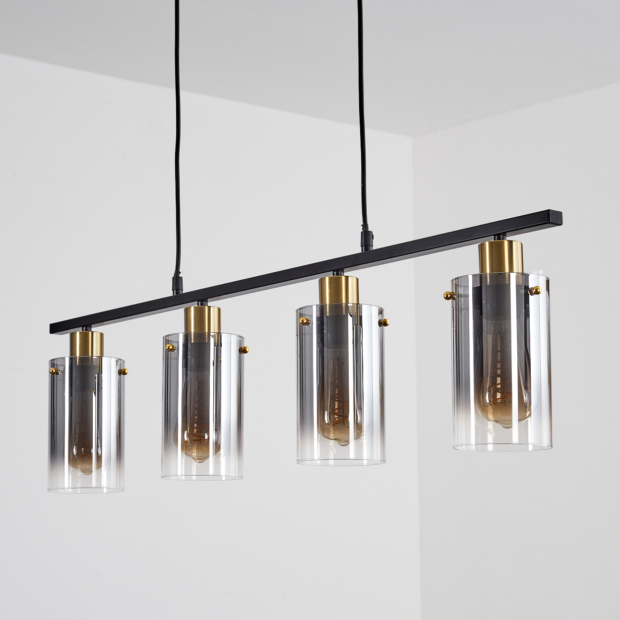 Eine moderne Deckenleuchte mit vier zylindrischen Glasschirmen, schwarzem Metallrahmen und goldenen Akzenten, an einer weißen Wand.