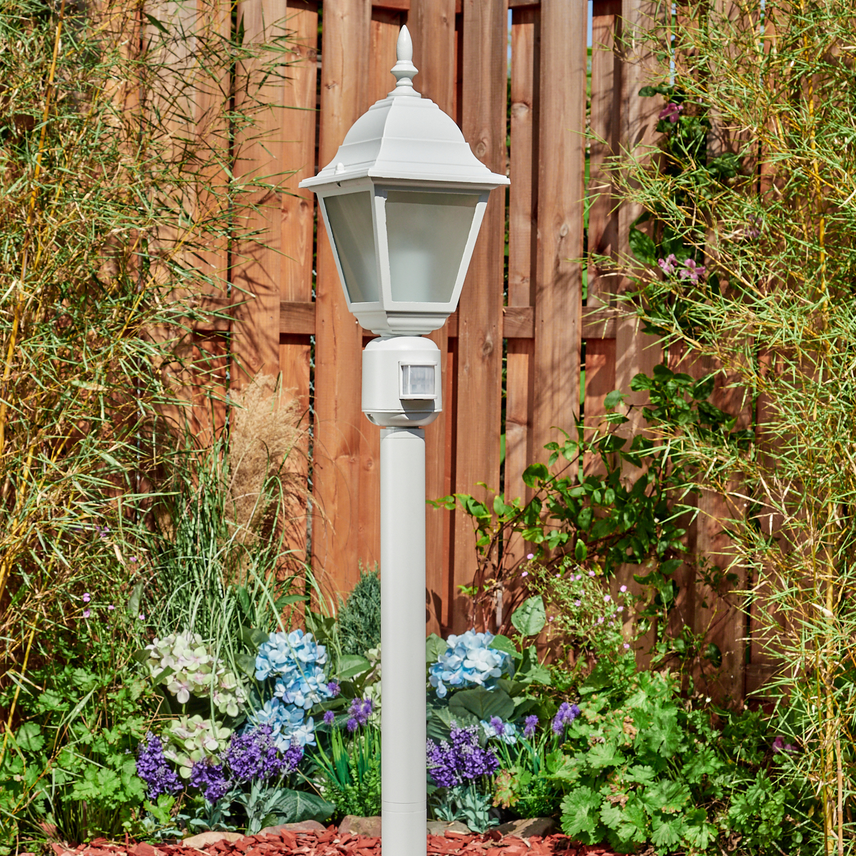 Eine weiße Außenlampe steht in einem Garten mit blauen und lila Blumen. Im Hintergrund befinden sich ein Holzzaun und Bambus.