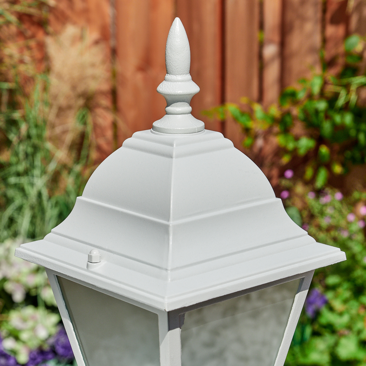 Eine weiße Gartenlampe mit spitzer Spitze steht vor einem Holzzaun und Grün.