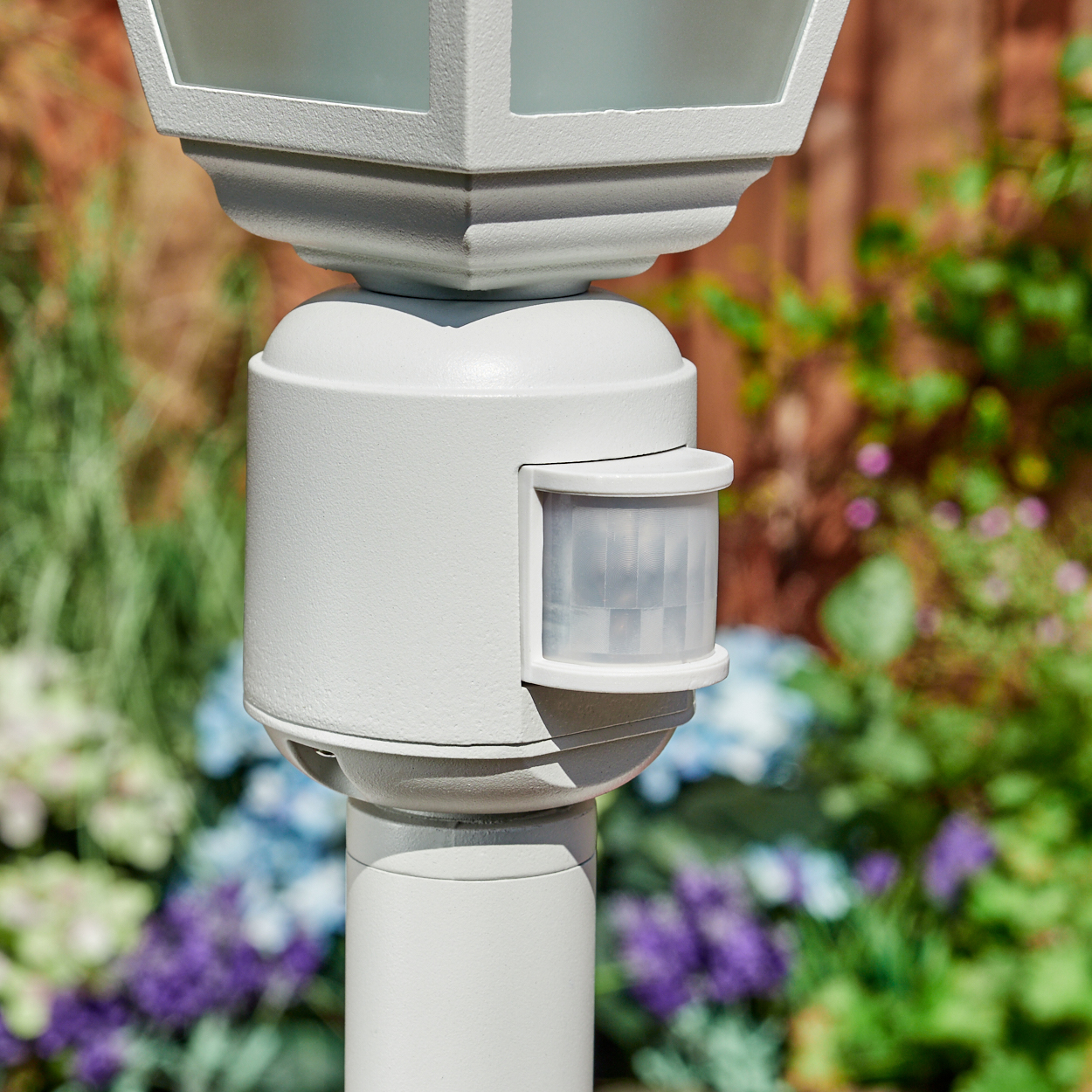 Eine weiße Außenlaterne mit Bewegungsmelder. Der Hintergrund umfasst verschwommenes Grün und Blumen mit einem Holzzaun.