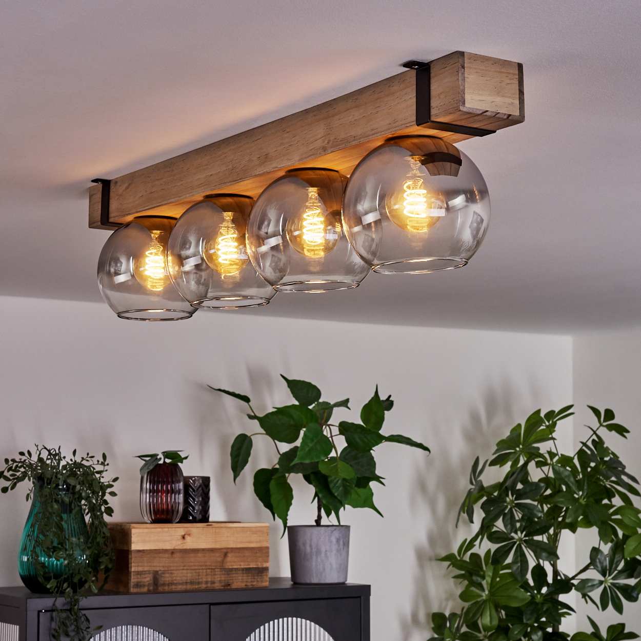 Ein Holzbalken mit fünf Glaskugelleuchten hängt von der Decke. Darunter stehen Pflanzen und Dekorationsartikel auf einem Schrank.