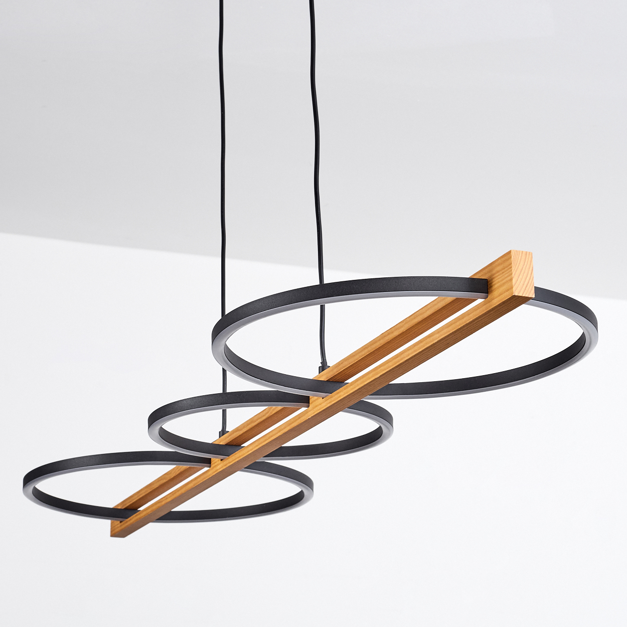 Moderne Hängelampe mit drei schwarzen kreisförmigen Lichtern und Holzbalken. Der Hintergrund ist eine schlichte weiße Wand und Decke.