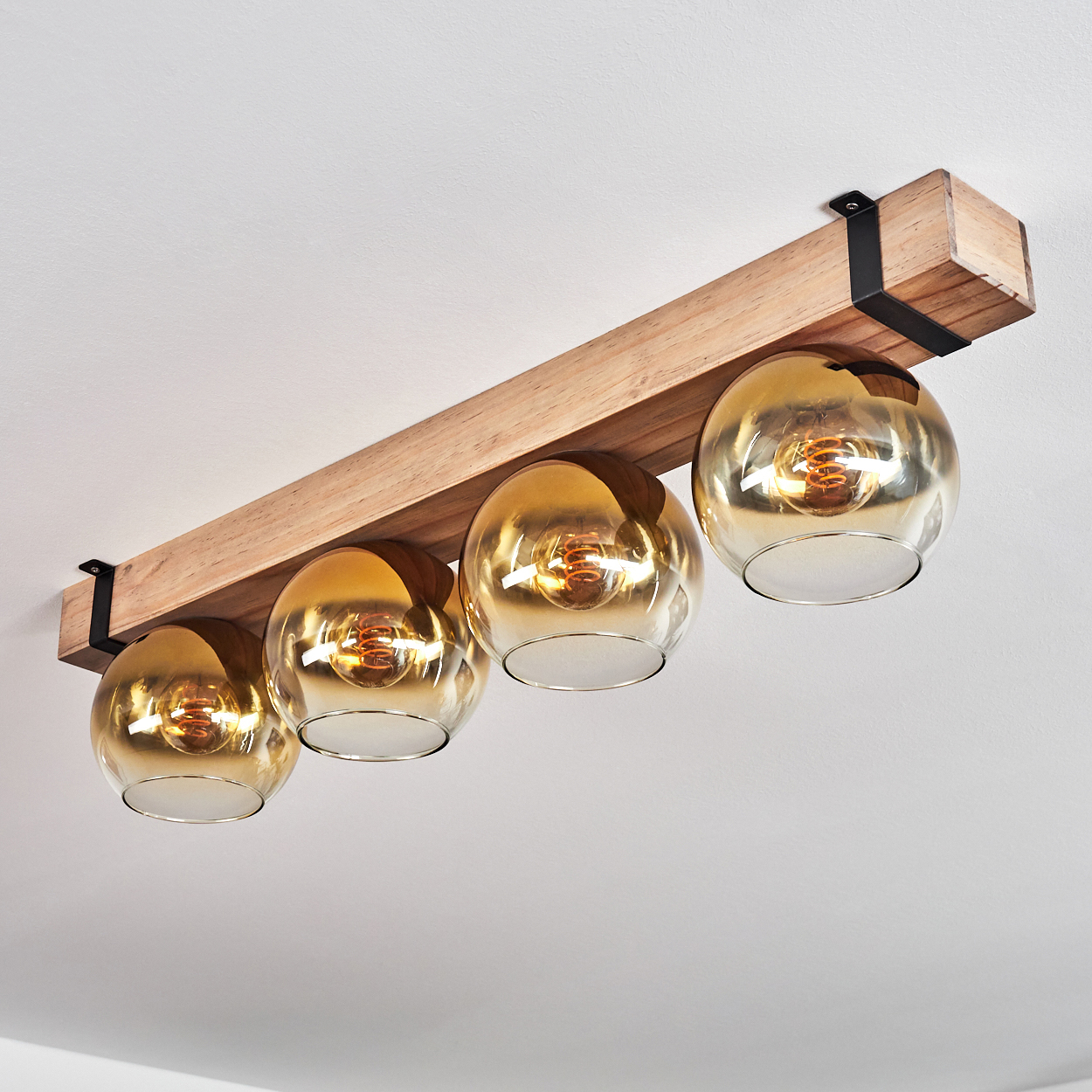 Eine Deckenleuchte aus Holzbalken mit vier goldfarbenen Glasschirmen. Schwarze Metallhalterungen stützen den Balken an einer weißen Decke.