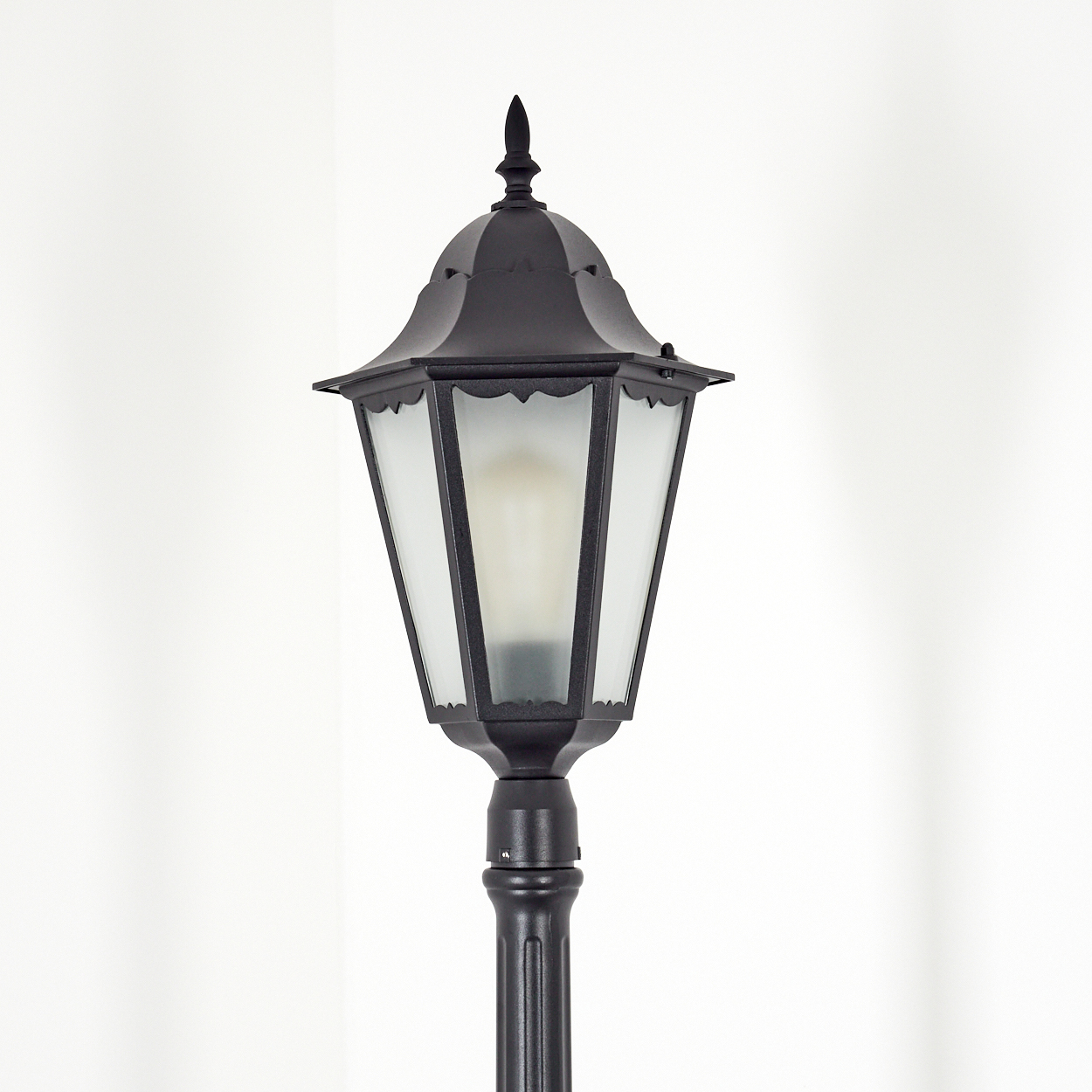 Schwarze Außenlaterne mit Glasscheiben und dekorativer Spitze, vor weißem Hintergrund.