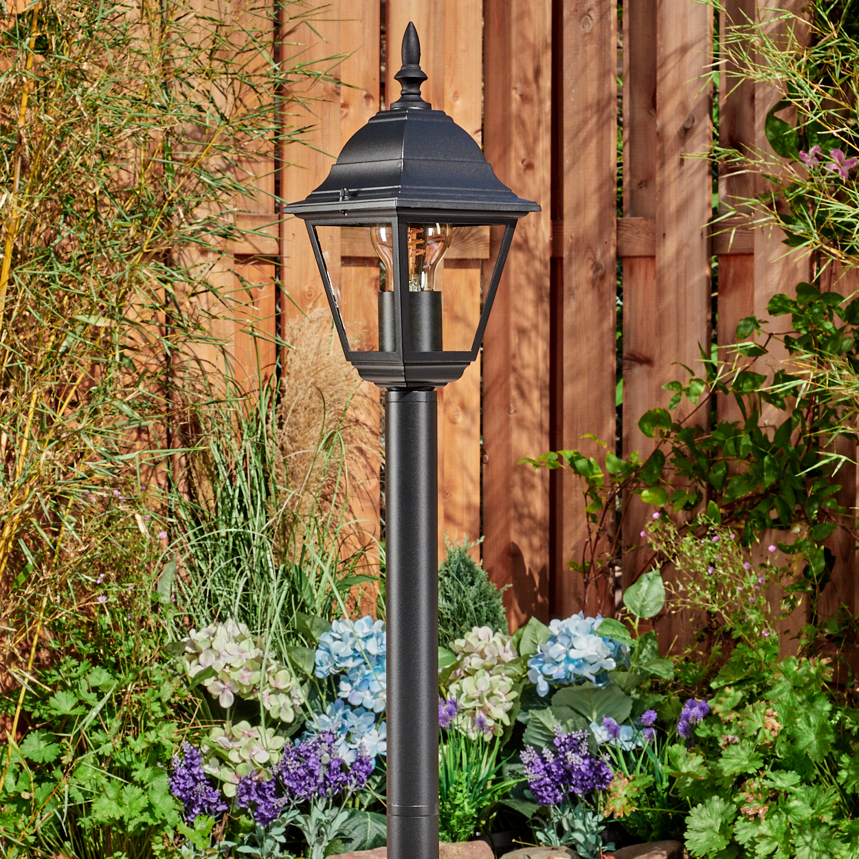 Schwarze Laterne im Garten, Blumen im Vordergrund, Holzzaun im Hintergrund. Die Lampe hat eine Glühbirne.