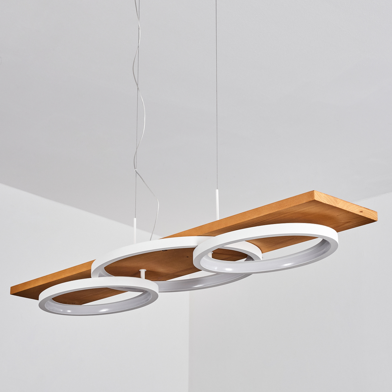 Eine moderne Leuchte. Sie hat ein Holzbrett mit drei weißen kreisförmigen Lichtern, die von der Decke hängen, vor weißem Hintergrund.