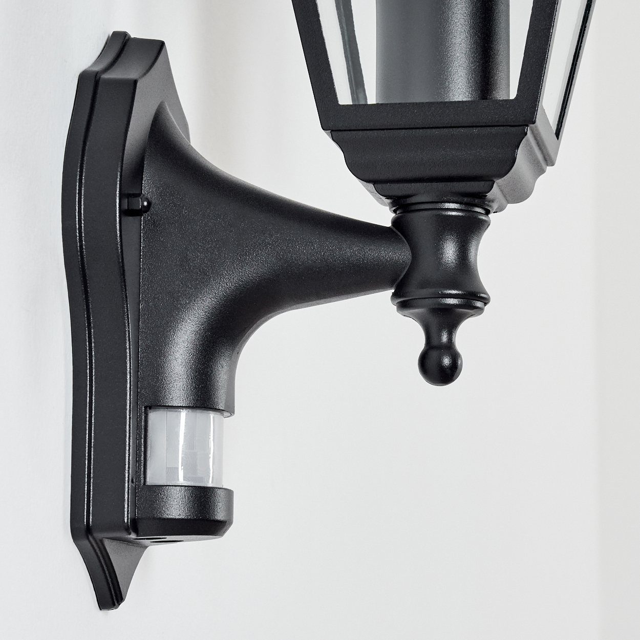 Schwarze Außenlampe mit Bewegungssensor, an einer weißen Wand montiert. Die Lampe hat ein klassisches Design.