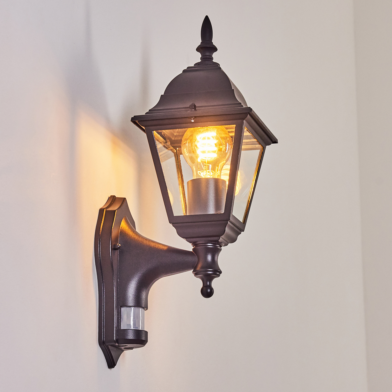 Eine schwarze Außenlampe mit leuchtender Glühbirne ist an einer weißen Wand befestigt. Sie hat einen Bewegungssensor.
